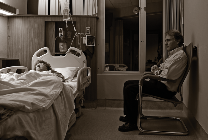 End-of-Life Care. Woman in hospital bed with man seated in chair