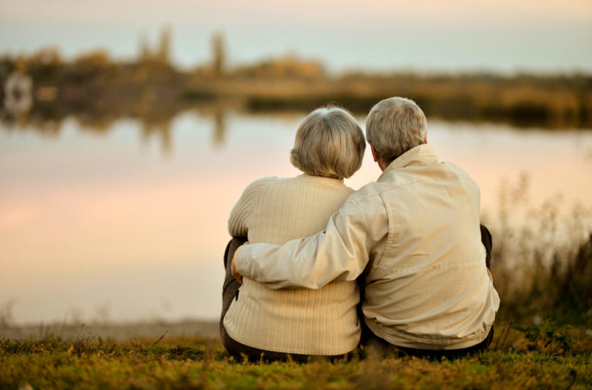 Shot from behind of male and female elderly couple sitting on ground and looking out at lake