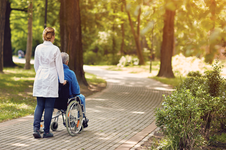 nurse pushing elderly man in a wheel chair down brick path in the woods