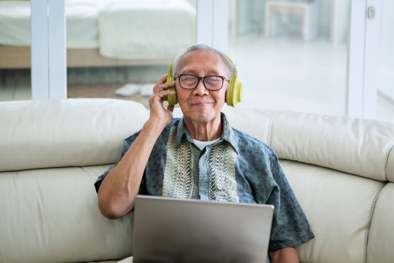 Senior listening to music with headphones and laptop computer on the sofa.