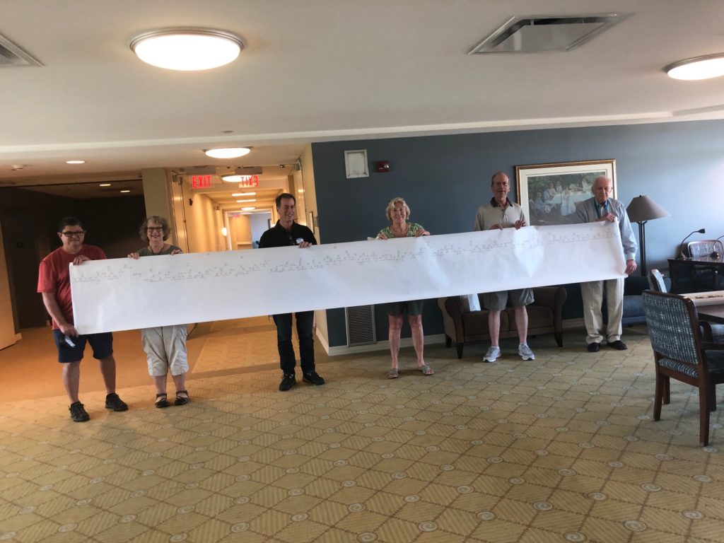 Six people stand in a line holding a giant chart of a family tree on white paper.