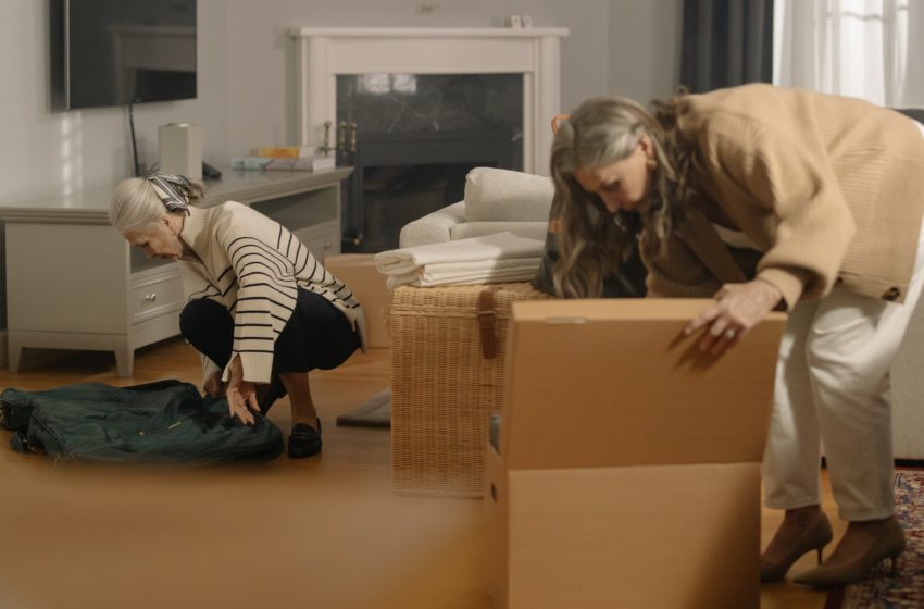 Two women, one with white hair pulled back and one with long gray hair, pack moving boxes.