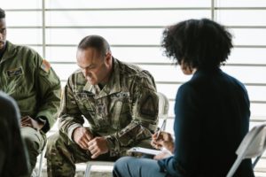 A group of U.S. Army veterans sit in a circle. One is looking down, his elbows on his knees, looking serious.