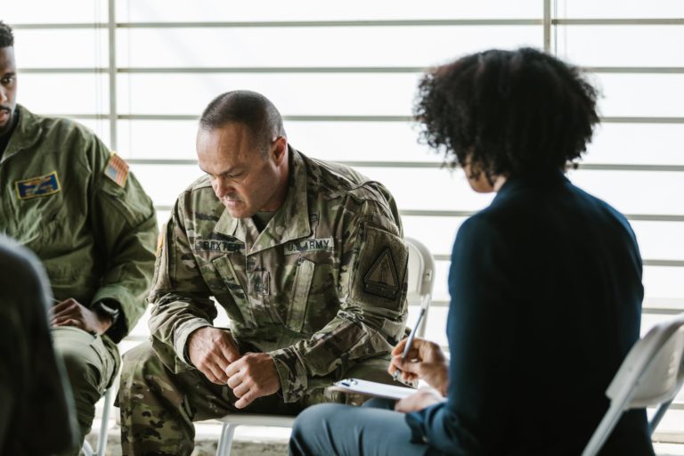 A group of U.S. Army veterans sit in a circle. One is looking down, his elbows on his knees, looking serious.