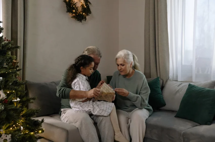 An older couple sits with their grandchild on a couch, near a Christmas tree.