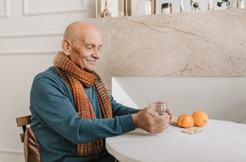 Old man, bald, wearing a sweater and scarf sits at a table with oranges.