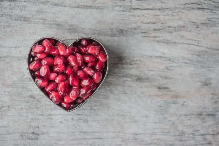 Pomegranate seeds in a silver, heart-shaped bowl, shot from above, set on a gray-tinted wooden table