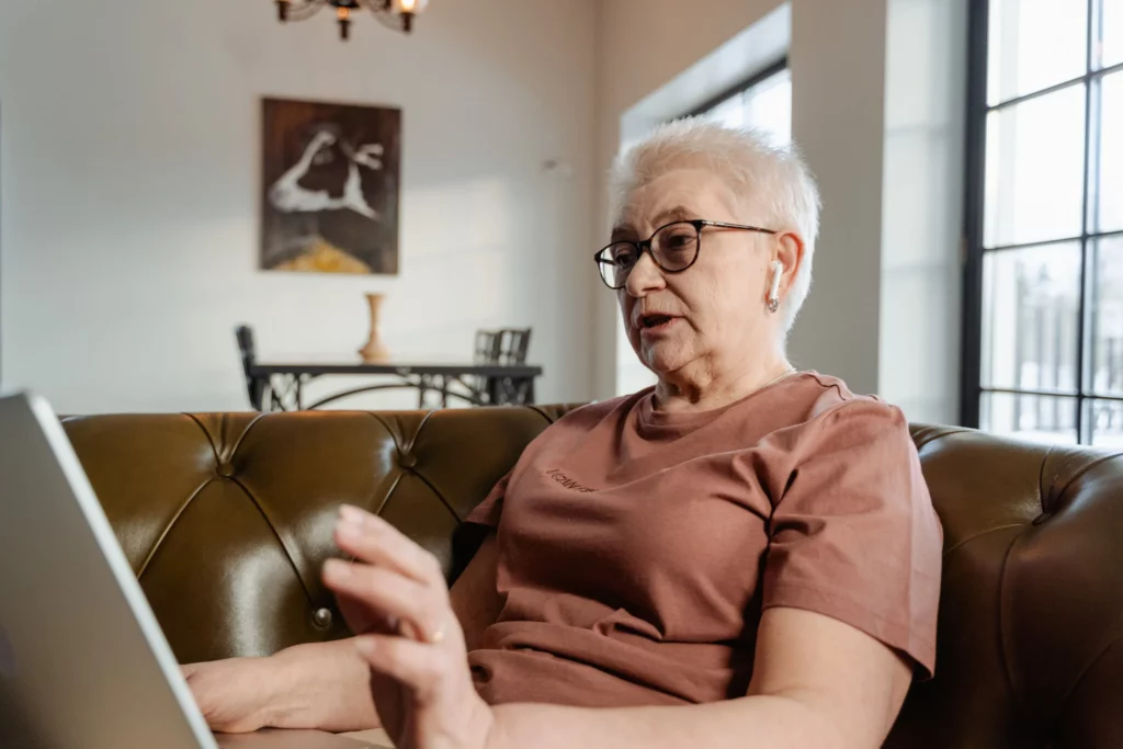 Older woman with short white hair, wearing a clay-colored tshirt, looks at a laptop screen.