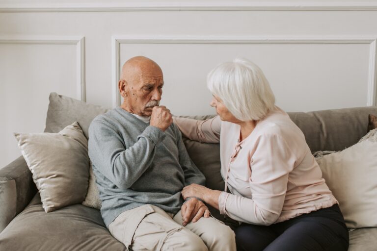 An older couple sits on a couch, talking. The man is in a blue/gray sweatshirt and coughing.