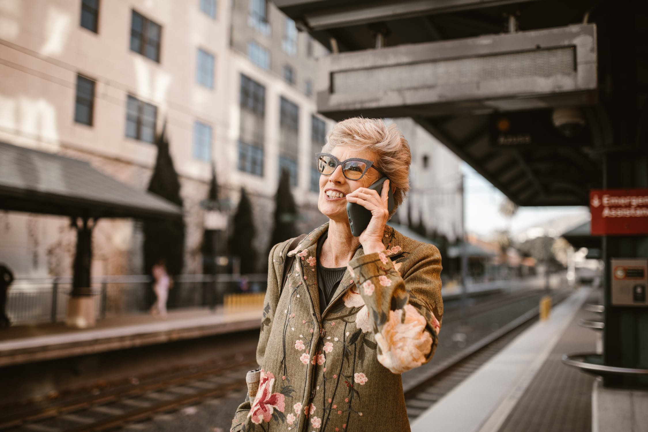 A woman with blonde hair, wearing a flowered jacket, smiles as she talks on the phone while walking along train tracks at a train station.