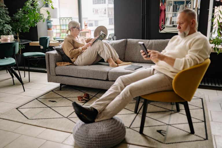 An older couple reads in their living room, one sitting on the couch with a book, and the other in a chair in the foreground, reading from an e-reader