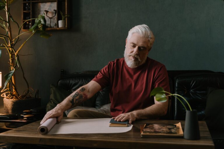 An older man with white hair wearing a maroon t-shirt sits at a table. On his arm is a tattoo that says DANGER