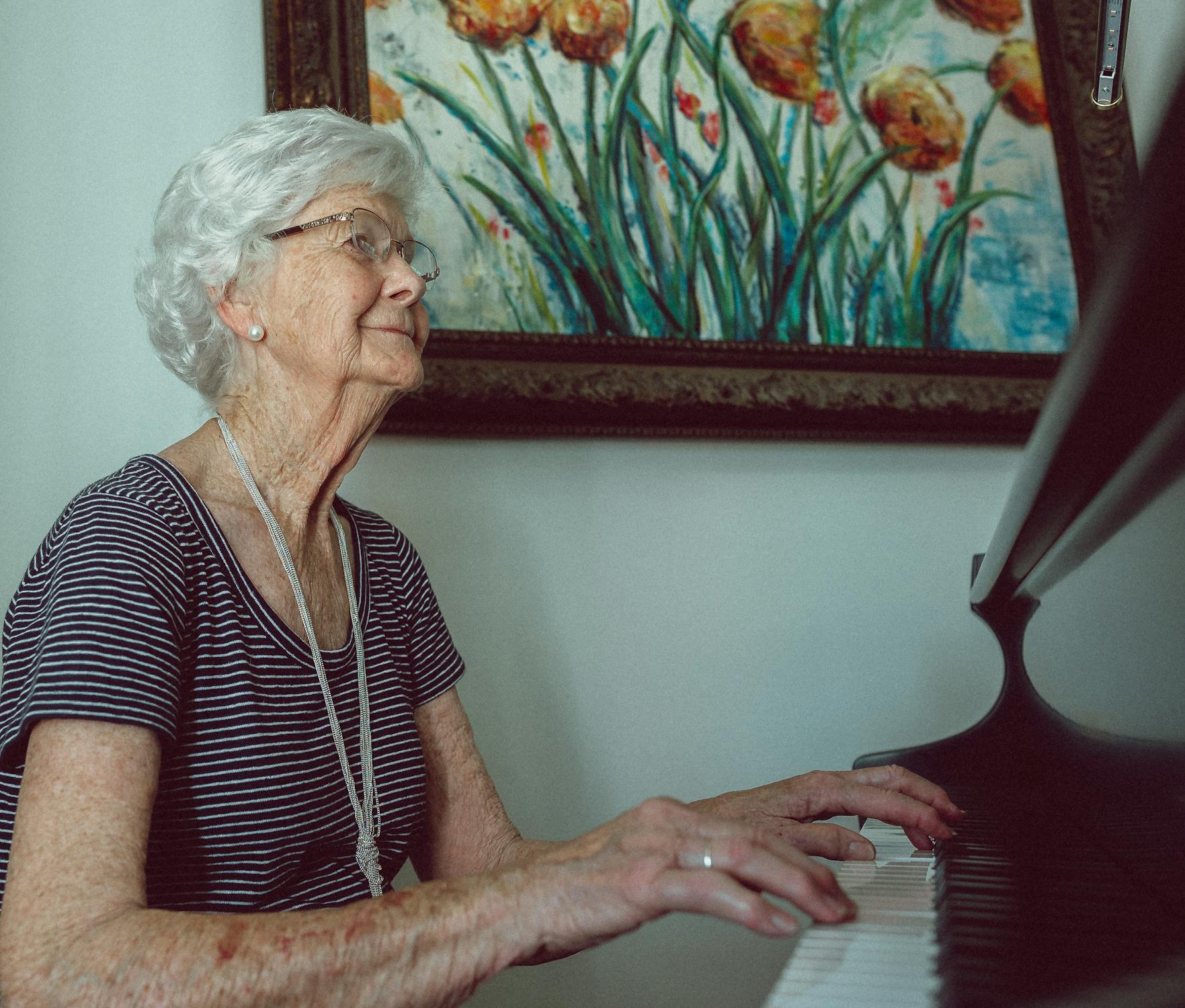 Older woman with white hair, wearing a purple shirt, plays the piano.