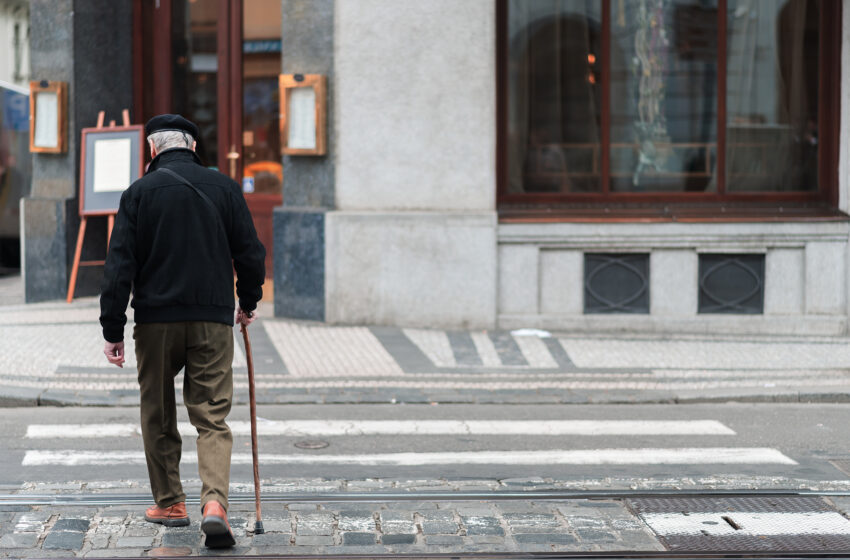 A man with gray hair walks across a city street, using a cane to steady himself.