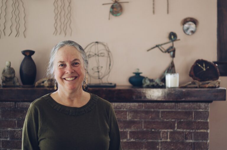 Anthropologist Rebecca Berman, PhD, stands before a brick fireplace. She's wearing a green knit shirt, and her gray hair is pulled back into a ponytail. She's smiling.