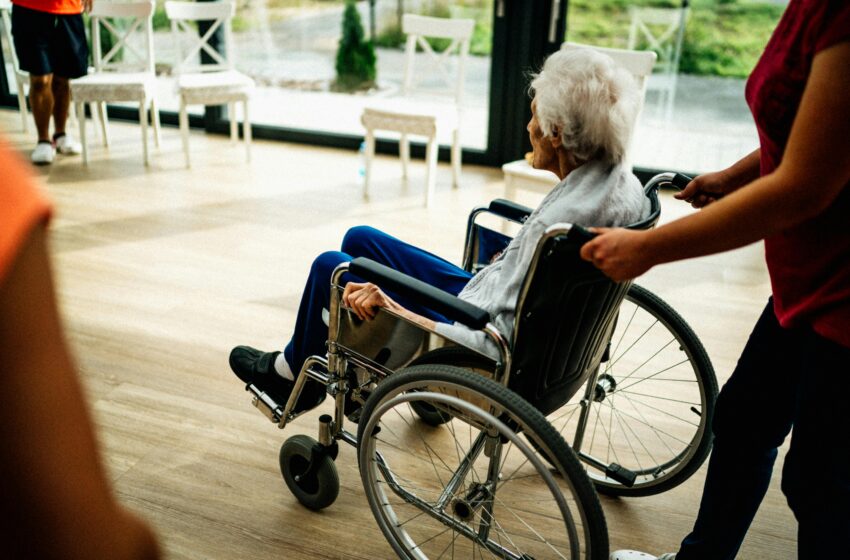 A person (face not visible) is pushing an older lady with white hair in a wheelchair