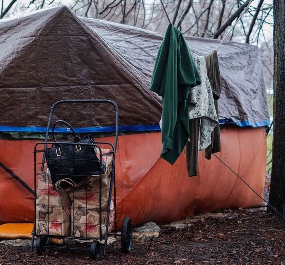 Clothes hang outside an orange tent. The tent is under a tree, and there's a shopping cart beside it filled with grocery bags and a black purse.