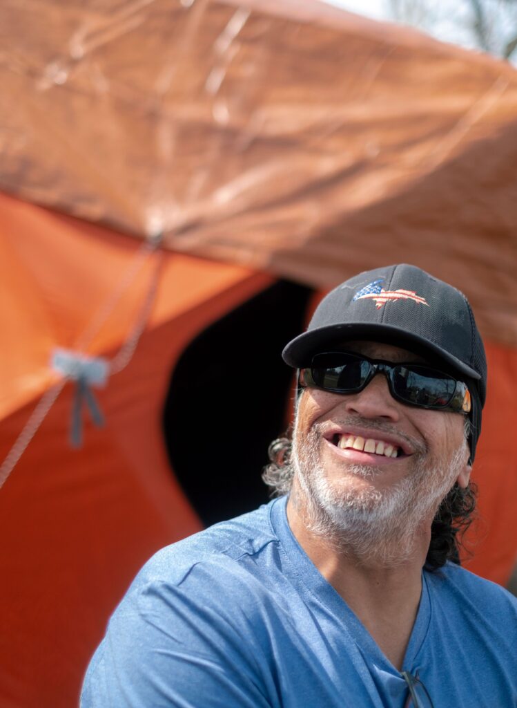 A man sits before an orange tent, wearing a blue shirt, sunglasses, and a baseball hat. He's smiling upward at something just out of frame.