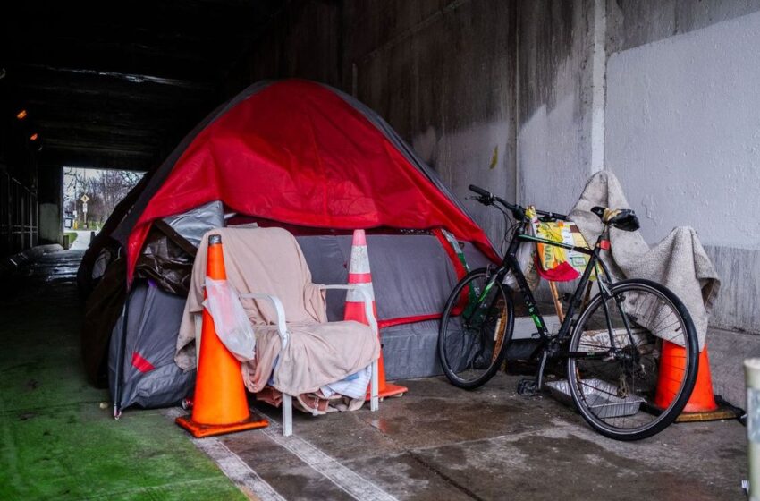 A red tarp covers a gray tent that has been set up under an overpass. Orange cones and a bike stand outside of the tent.