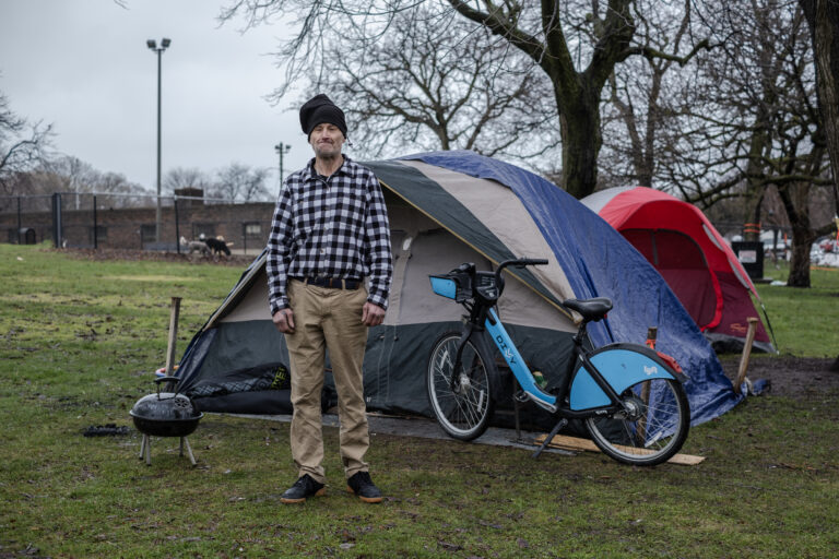 Scott Anthony Franklin stands outside a blue and gray tent. He's wearing a black-and-white checkered shirt, a black hat, and khaki pants.