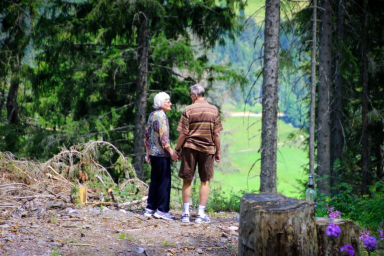 Two older adults stand in a forest together, looking out over a clearing.