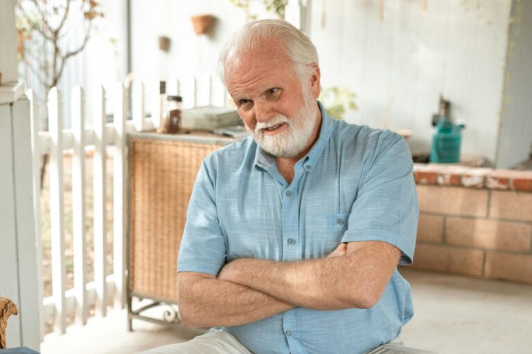 A man in a blue collared shirt crosses his arms. He has a white beard and is bald. He looks slightly frustrated.