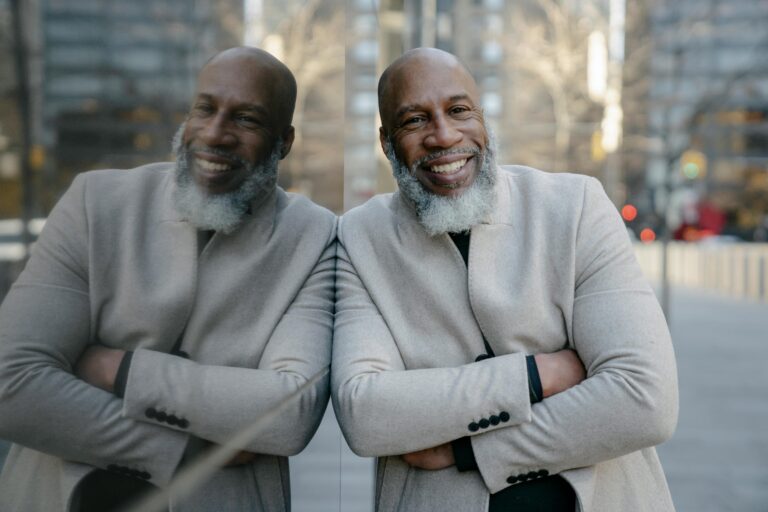 A man leans against a wall outdoors, smiling. He has his arms crossed and is wearing a light, wool winter coat