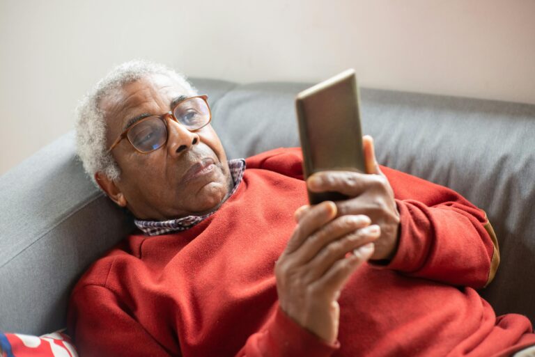 An older man reclines on a couch. He's wearing a red sweater, glasses, and looking at his cell phone.