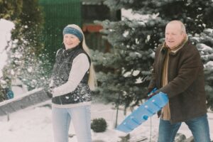 A man and woman stand side by side, each holding a shovel and throwing snow in the air.