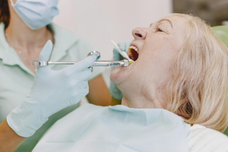 An older woman with light hair holds he mouth open as a dentist inspects her teeth.