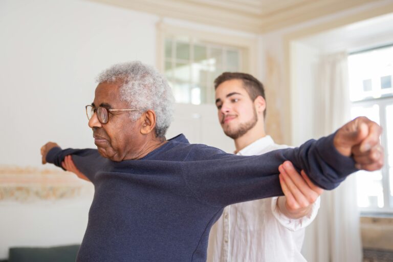 An older man stretches his arms wide, with the help of a physical therapist.