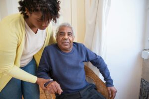A younger woman in a light yellow sweater helps an older man in a blue shirt get into a chair.