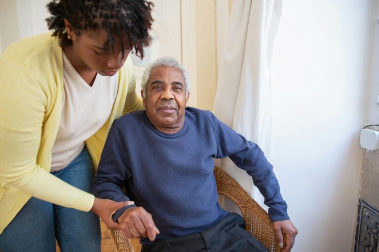 A younger woman in a light yellow sweater helps an older man in a blue shirt get into a chair.