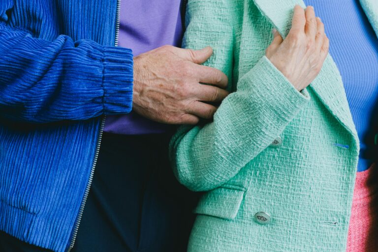 An older man in a blue jacket and purple shirt holds onto the arm of an older woman in a teal jacket and blue shirt. We don't see their faces — only a close up of their arms together.