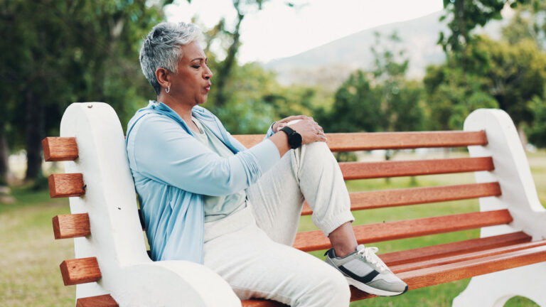 Older woman sitting on a park bench holding her knee. Mobility issues, knee pain.