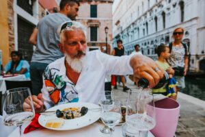 A bearded man with white hair sits at an outdoor cafe in Italy, pouring wine