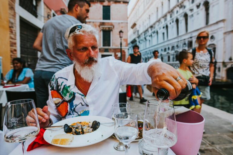 A bearded man with white hair sits at an outdoor cafe in Italy, pouring wine