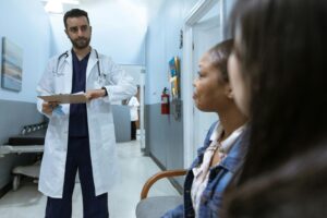 A doctor stands in a hallway, looking at two patients seated and waiting