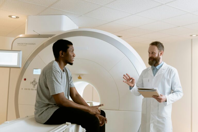 A man sits on the bed of a CT scan machine, talking to a doctor.