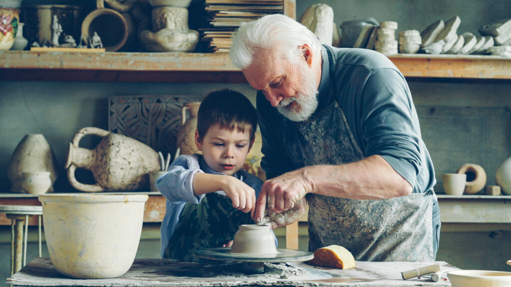 A senior man teaches a child how to make a piece of pottery on a pottery wheel